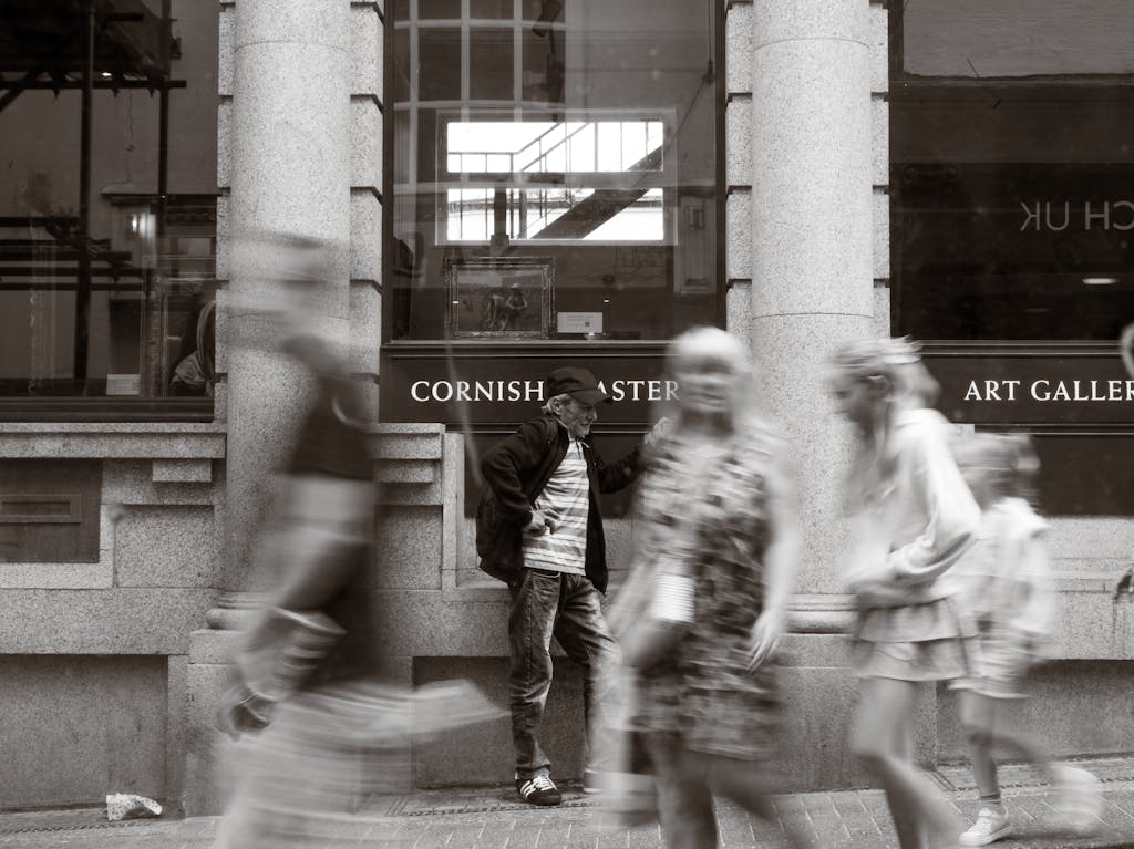 Man standing against art gallery as blurred pedestrians pass by in grayscale.